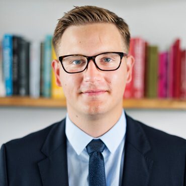 A man in glasses and suit standing next to books.